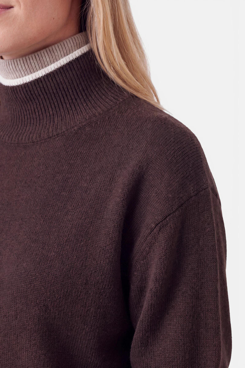 Eine Person, die einen dunkelbraunen Pullover aus einer Woll-Kaschmir-Mischung mit Stehkragen und einem mehrlagigen Kragen mit weiß-beiger Streifenborte trägt.