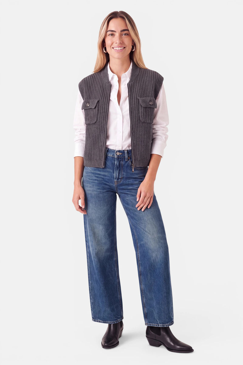 Eine lächelnde Frau, die eine graue, gerippte Strickweste mit Stehkragen über einem weißen Hemd trägt, dazu eine blaue Jeans mit weitem Bein und schwarze Stiefel.