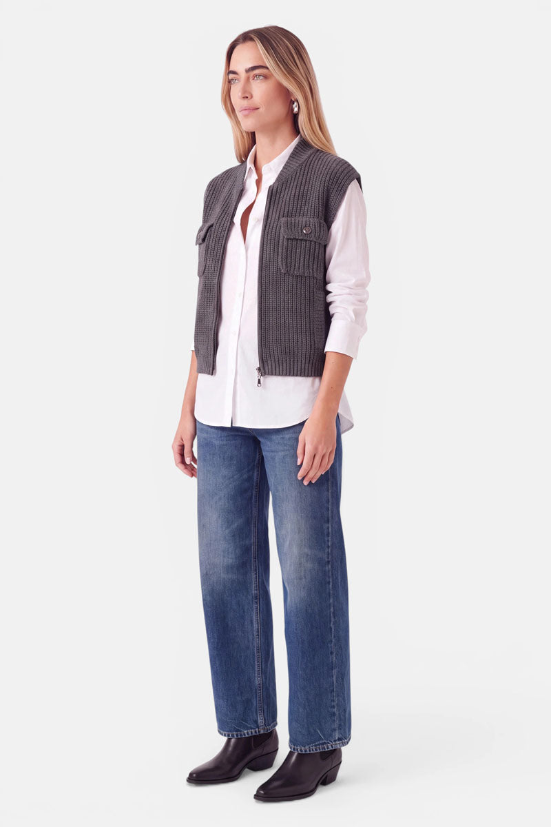 Eine Frau trägt eine graue Wolle-Kaschmir-Weste mit Reißverschluss über einem weißen Hemd und einer blauen Jeans mit geradem Bein, dazu schwarze Stiefel.