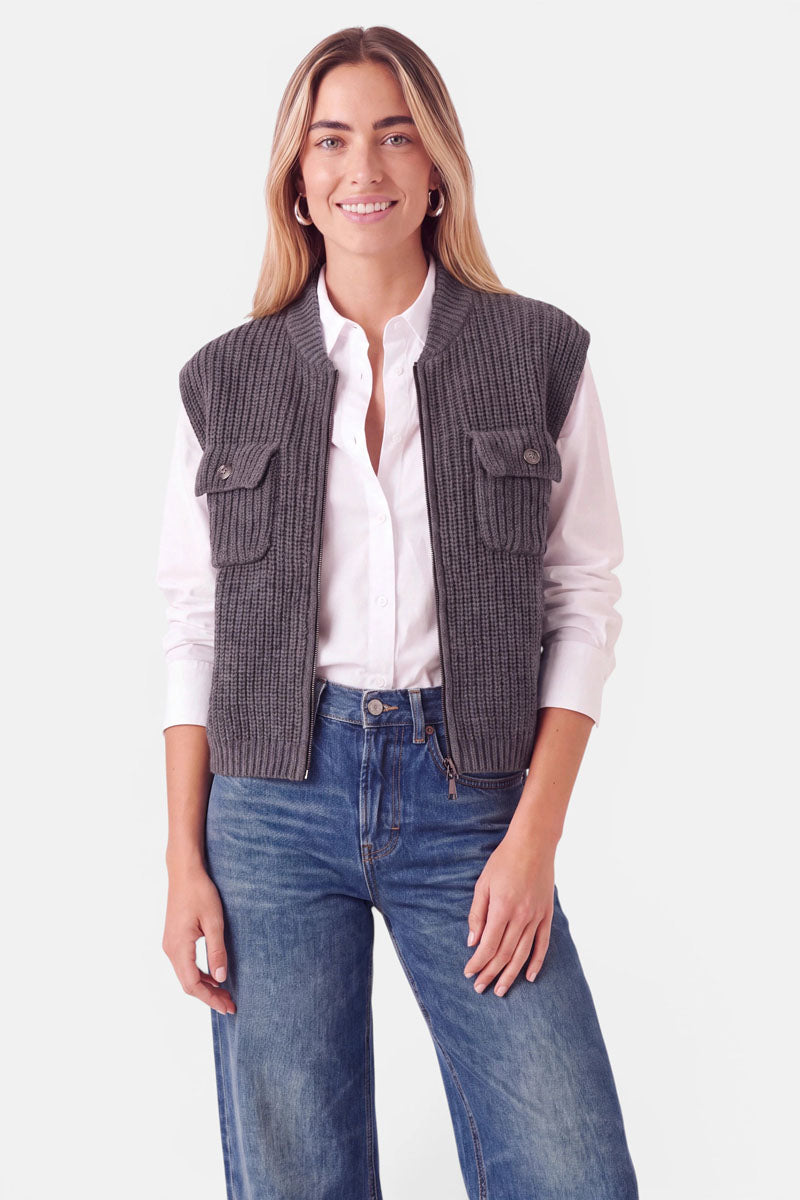 Eine lächelnde Frau trägt eine dunkelgraue, gerippte Strickweste aus Wolle und Kaschmir mit Stehkragen und zwei geknöpften Brusttaschen über einem weißen Hemd und einer blauen Jeans.