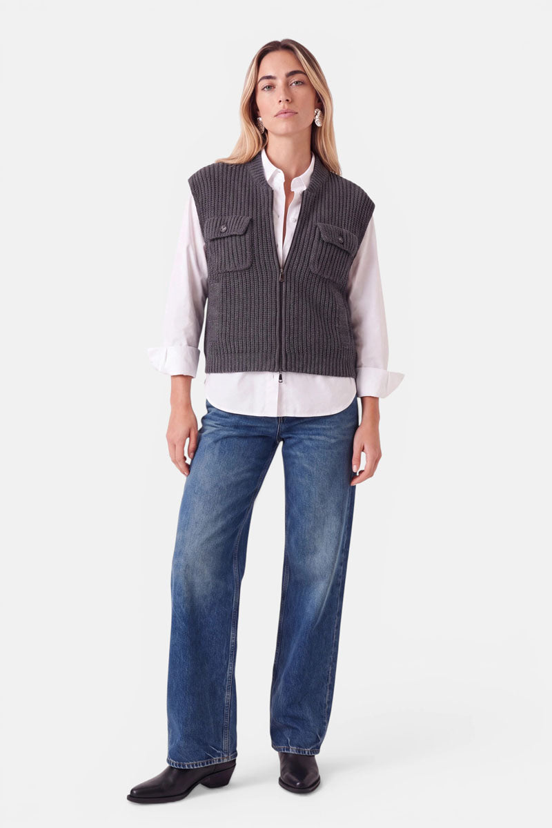 Eine Frau trägt eine graue Wolle-Kaschmir-Weste mit Reißverschluss über einem weißen Hemd und einer blauen Jeans. Außerdem trägt sie schwarze Stiefel.