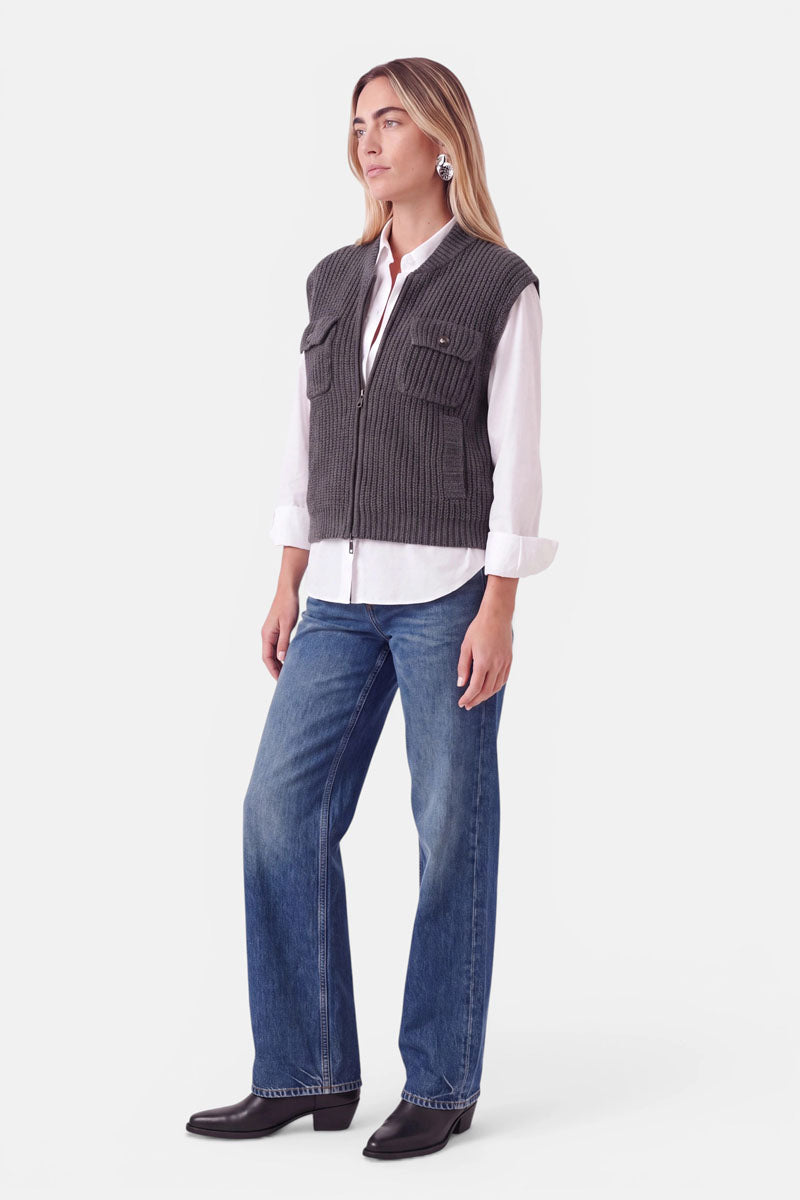 Eine Frau trägt eine graue Wolle-Kaschmir-Weste mit Reißverschluss über einem weißen Hemd und einer blauen Jeans. Sie steht da, blickt zur Seite und trägt schwarze Stiefel.
