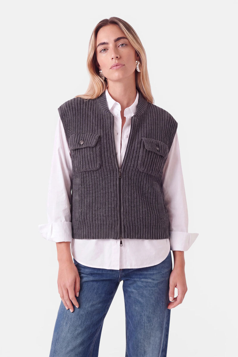 Eine Frau trägt eine anthrazitfarbene Strickweste aus einer Woll-Kaschmir-Mischung mit Stehkragen über einem weißen Hemd und einer blauen Jeans. Die Weste mit Reißverschluss verfügt über zwei auffällige Brusttaschen mit Knopfklappen.
