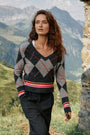 Eine Frau mit welligem braunem Haar trägt einen Strickpullover mit einem Argyle-Muster in Rot, Grau und Schwarz, dazu eine Nadelstreifenhose. Sie spaziert im Freien durch eine bergige Graslandschaft, wobei rechts teilweise Steinruinen zu sehen sind und im Hintergrund Hügel und Berge liegen.