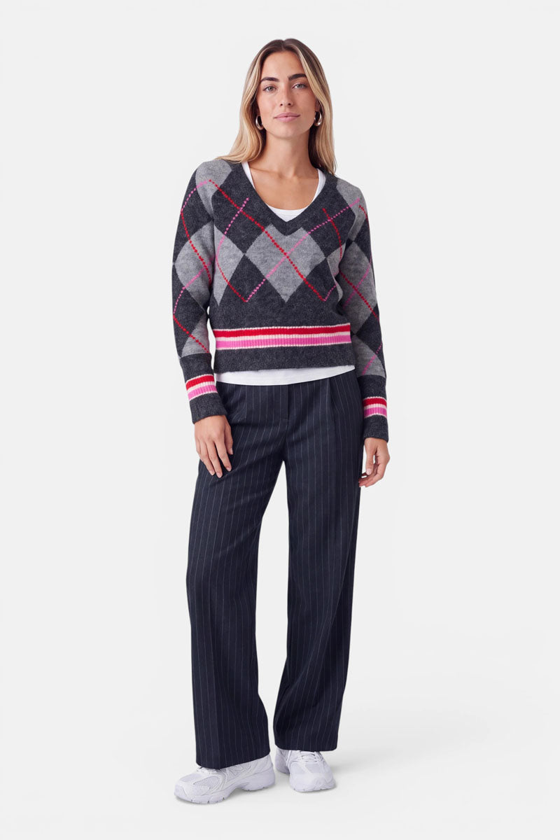 Eine Frau steht vor einem weißen Hintergrund und trägt einen grauen Strickpullover mit Argyle-Muster und Merinoanteil sowie roten und weißen Akzenten, darunter ein weißes Hemd, eine dunkle, breit geschnittene Nadelstreifenhose und weiße Sneaker. Sie hat langes blondes Haar und einen neutralen Gesichtsausdruck, die Arme liegen entspannt an den Seiten.
