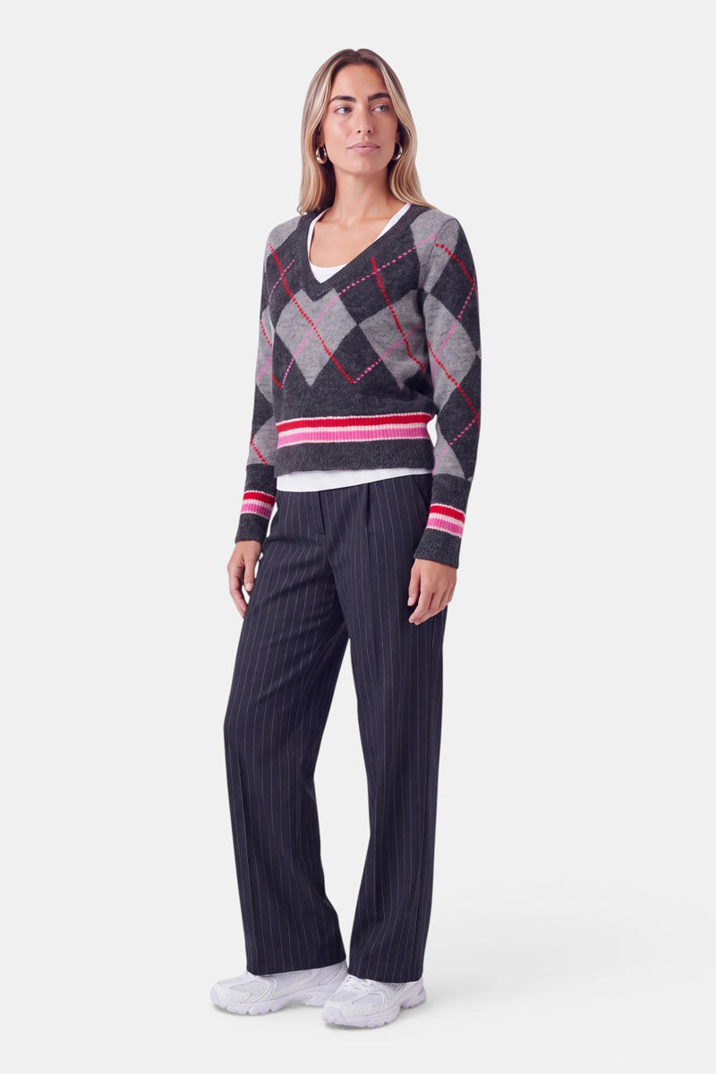 Eine Frau steht vor einem weißen Hintergrund und trägt einen grauen Strickpullover mit roten und weißen Akzenten im Argyle-Muster und einem hohen Merinoanteil, über einem weißen Hemd. Dazu trägt sie eine lockere schwarze Nadelstreifenhose und weiße Sneaker. Ihr Haar trägt sie offen, und sie blickt mit neutralem Gesichtsausdruck leicht zur Seite.