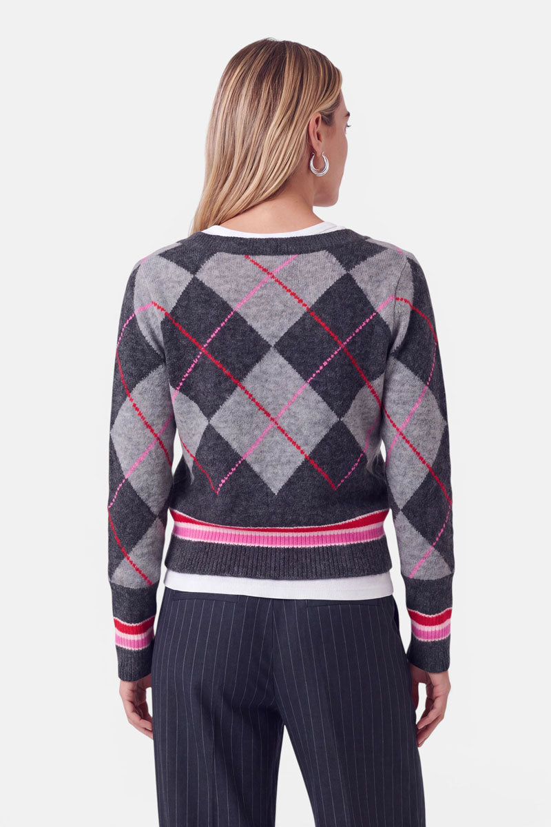 Eine Frau mit langen, glatten, hellbraunen Haaren, die silberne Creolen und einen grau-schwarzen Strickpullover mit Argyle-Muster und roten Akzenten trägt, dazu eine schwarze Nadelstreifenhose, steht mit dem Rücken zum Betrachter vor einem weißen Hintergrund.