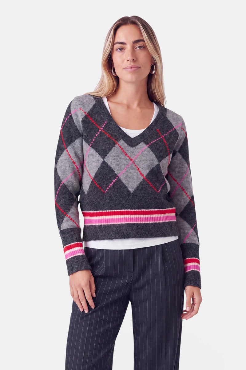 Eine Frau mit glattem blondem Haar trägt einen Strickpullover mit Merinoanteil, der ein grau-schwarz-rosa Argyle-Muster aufweist, über einem weißen Hemd und einer Nadelstreifenhose. Sie blickt mit neutralem Gesichtsausdruck nach vorne und steht vor einem schlichten weißen Hintergrund.