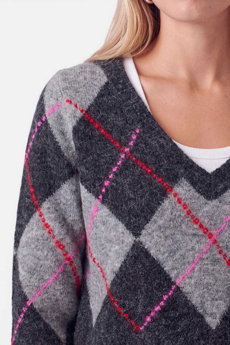 Eine Nahaufnahme einer Person, die einen flauschigen, grau-schwarzen Strickpullover mit Argyle-Muster und roten sowie rosa diagonalen Linien trägt. Die Person hat eine helle Haut und lange blonde Haare. Zu sehen sind nur die untere Hälfte des Gesichts, der Hals sowie ein Teil der Schulter und der Brust.