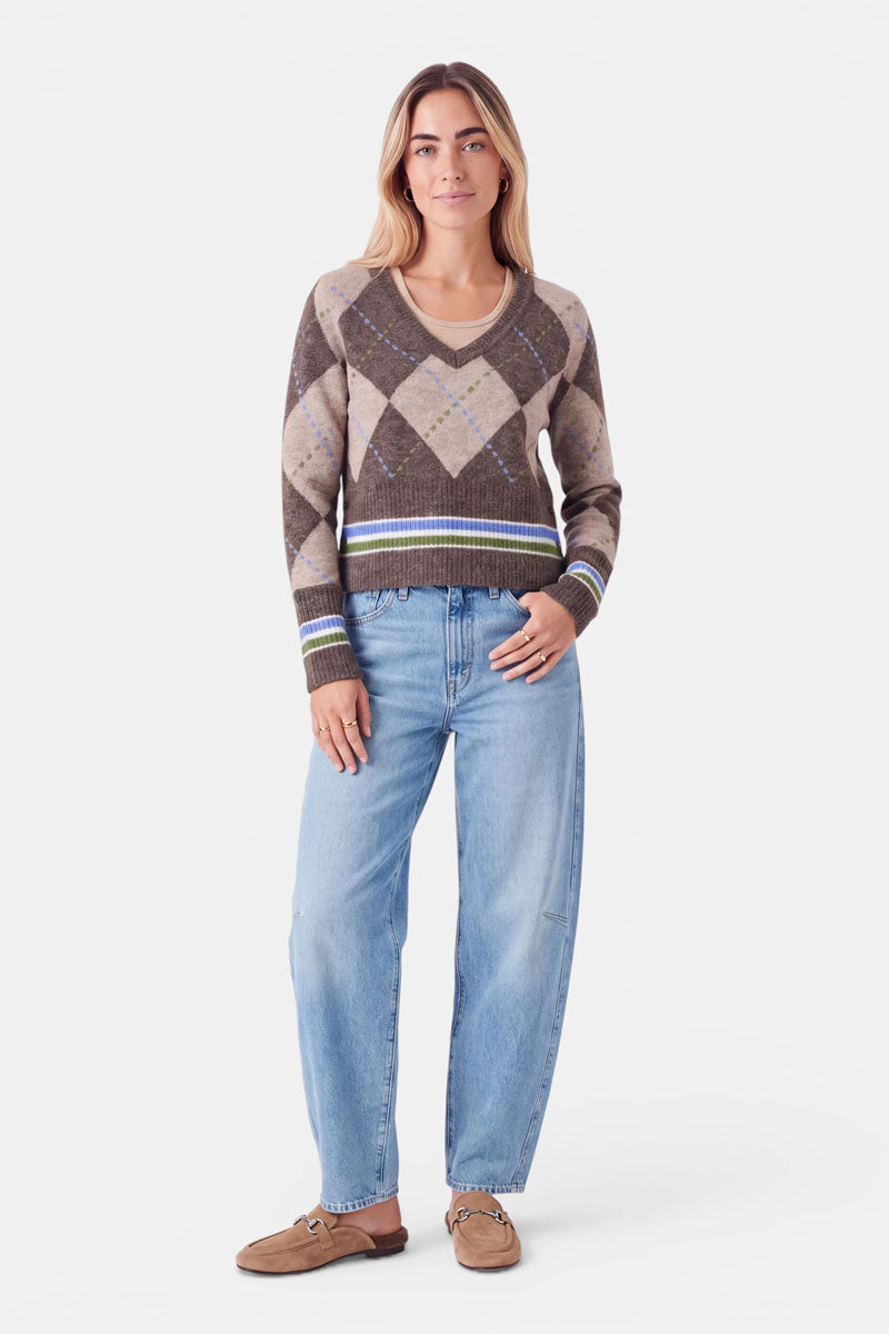 Eine Person mit langen, hellbraunen Haaren steht mit dem Blick nach vorne. Sie trägt einen braun-beigen Strickpullover mit grünem und blauem Argyle-Muster und Merinoanteil, hellblaue, locker sitzende Jeans und hellbraune Slipper. Der Hintergrund ist schlicht weiß. Ihre linke Hand ruht auf ihrem Oberschenkel.
