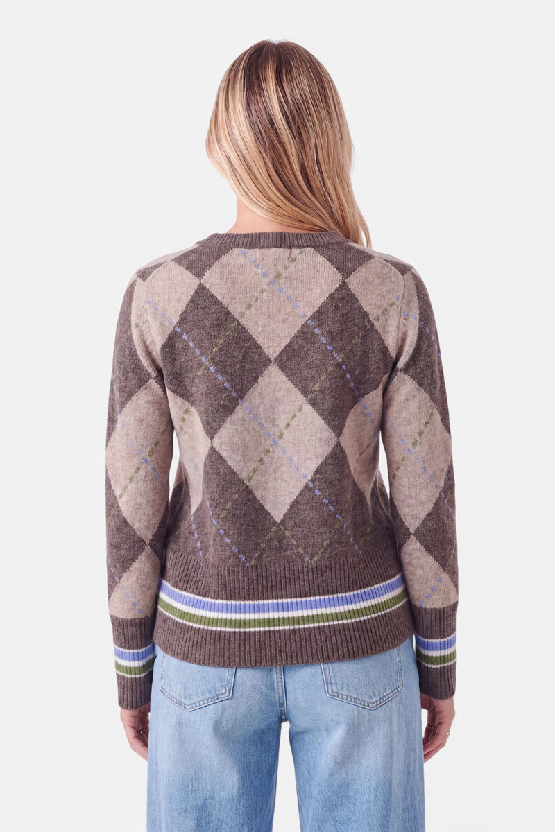 Eine Person mit langen, blonden Haaren trägt einen braun-beigen Strickpullover mit Argyle-Muster und grünen sowie blauen Details, dazu hellblaue Jeans. Sie steht mit dem Rücken zur Kamera. Der Pullover hat einen gerippten Saum, gerippte Ärmelbündchen sowie Streifen am Bund und an den Ärmeln.