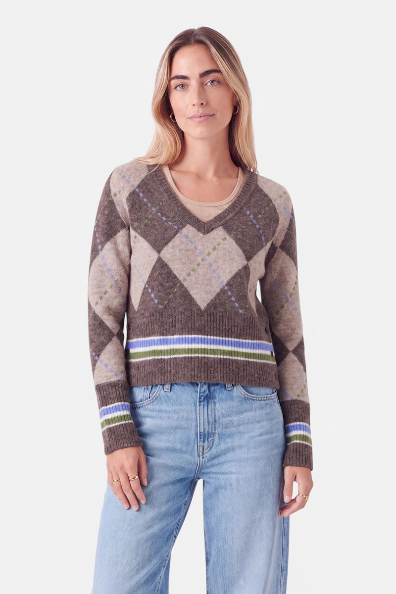 Eine Frau mit langen, hellbraunen Haaren trägt einen braunen Strickpullover im Argyle-Muster mit cremefarbenen, grünen und violetten Streifen an den Ärmelbündchen und am Saum. Sie steht mit dem Blick nach vorne, die Hände entspannt an den Seiten, in hellblauen Jeans vor einem schlichten weißen Hintergrund.