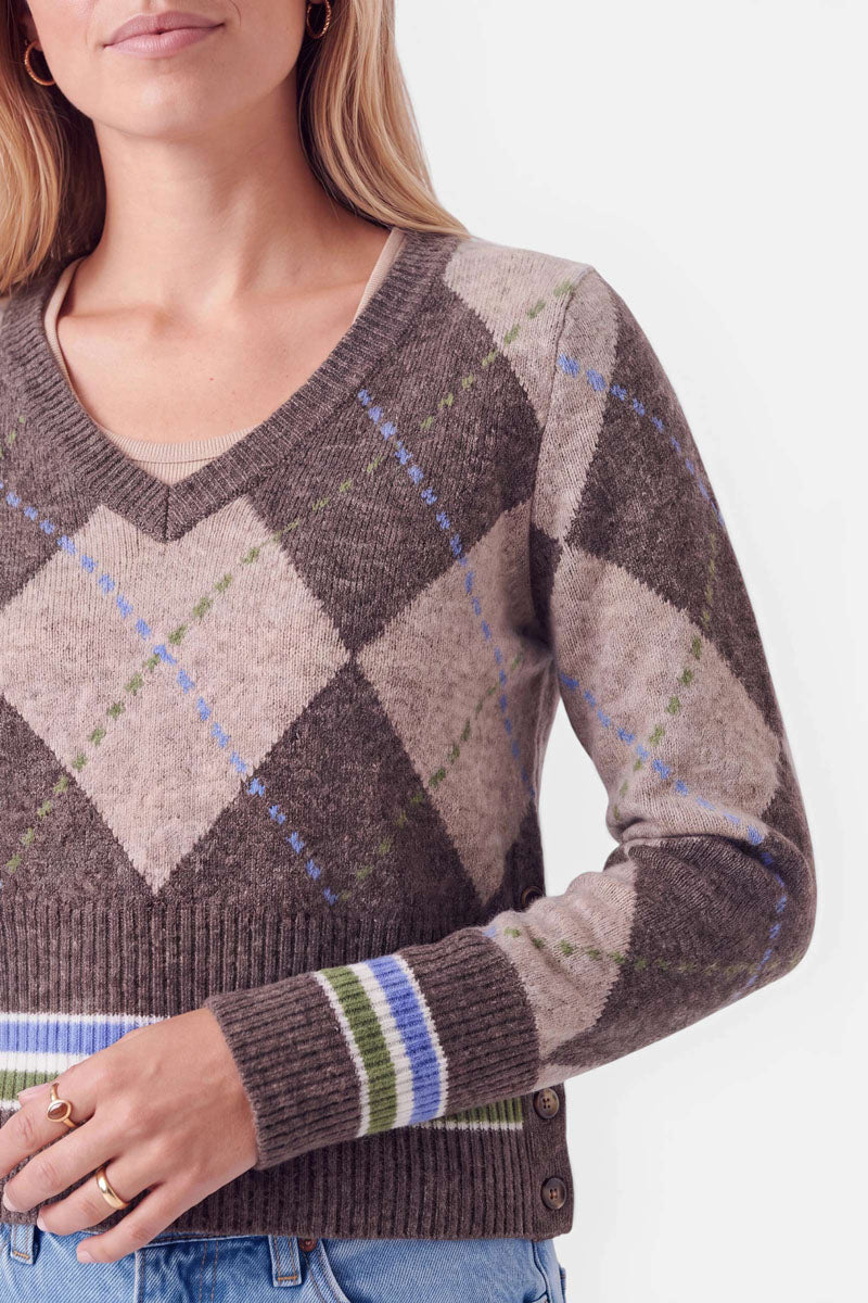 Eine Frau, die einen braun-beigen Strickpullover mit Argyle-Muster, blauen und grünen Akzenten, gerippten Bündchen und gestreiften Abschlüssen an den Ärmeln trägt. Der kuschelige Strickpullover enthält einen hohen Merinoanteil für zusätzlichen Tragekomfort. Außerdem trägt sie eine blaue Jeans und goldene Ringe. Ihr Gesicht ist teilweise zu sehen, und sie hat glattes, hellbraunes Haar.