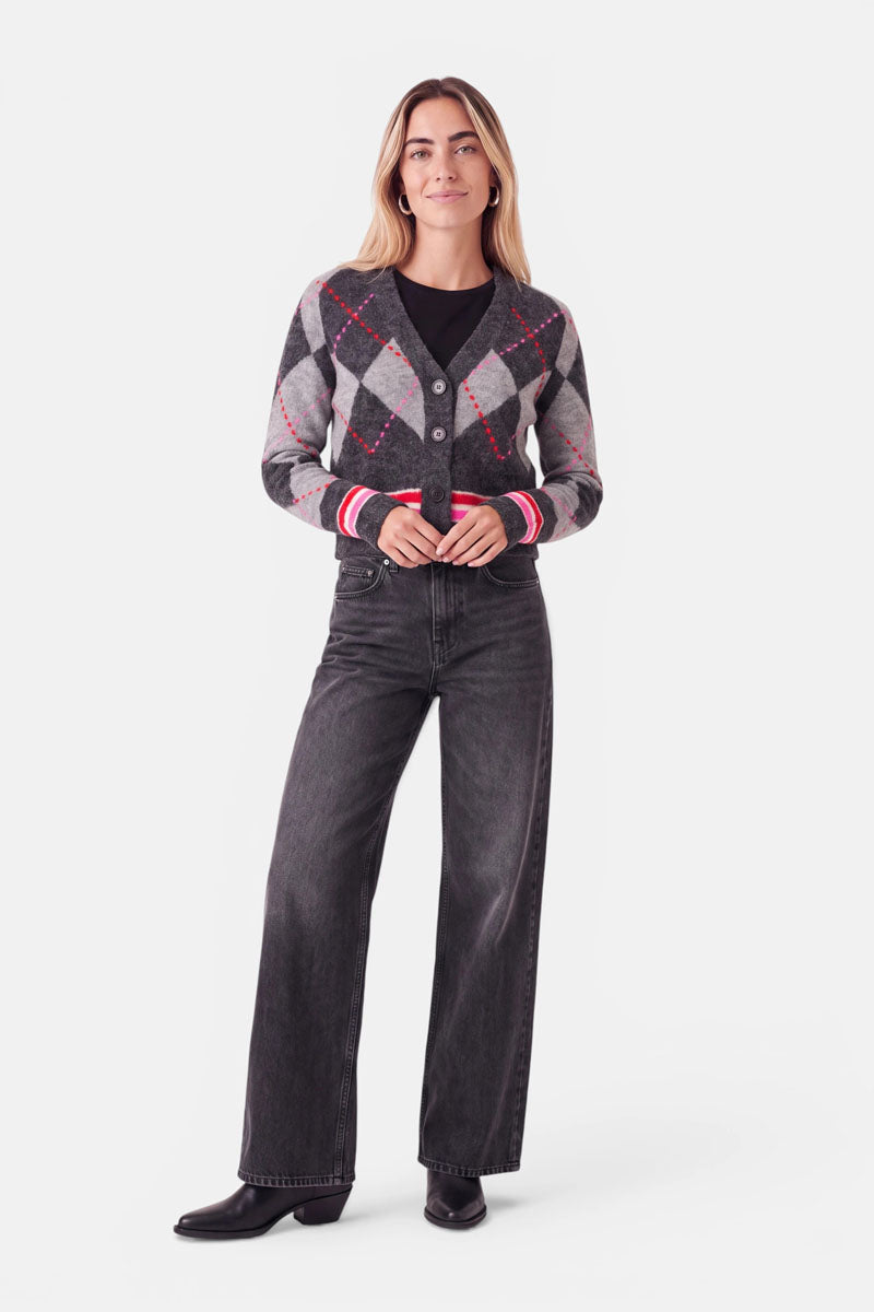 Eine Frau steht lächelnd da und trägt eine graue Strickjacke mit Argyle-Muster und roten sowie weißen Akzenten über einem schwarzen Shirt, dazu eine lockere dunkle Jeans und schwarze Schuhe. Sie hat langes, glattes Haar und steht vor einem schlichten weißen Hintergrund. Diese stilvolle Strickjacke enthält einen hohen Merino-Anteil für zusätzlichen Tragekomfort.