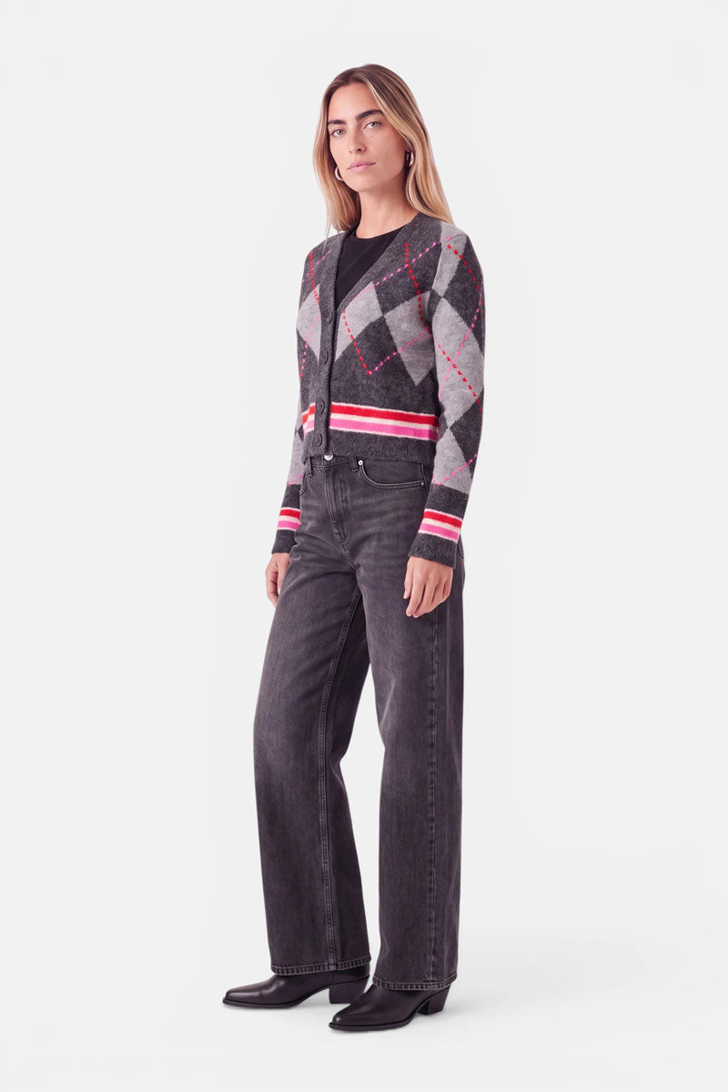 Eine Frau steht vor einem schlichten weißen Hintergrund und trägt eine graue Strickjacke mit Argyle-Muster und roten sowie weißen Akzenten, eine dunkle, hoch taillierte Jeans mit weitem Bein und schwarze Stiefel. Sie hat hellbraunes Haar, das in der Mitte gescheitelt ist, und einen neutralen Gesichtsausdruck.