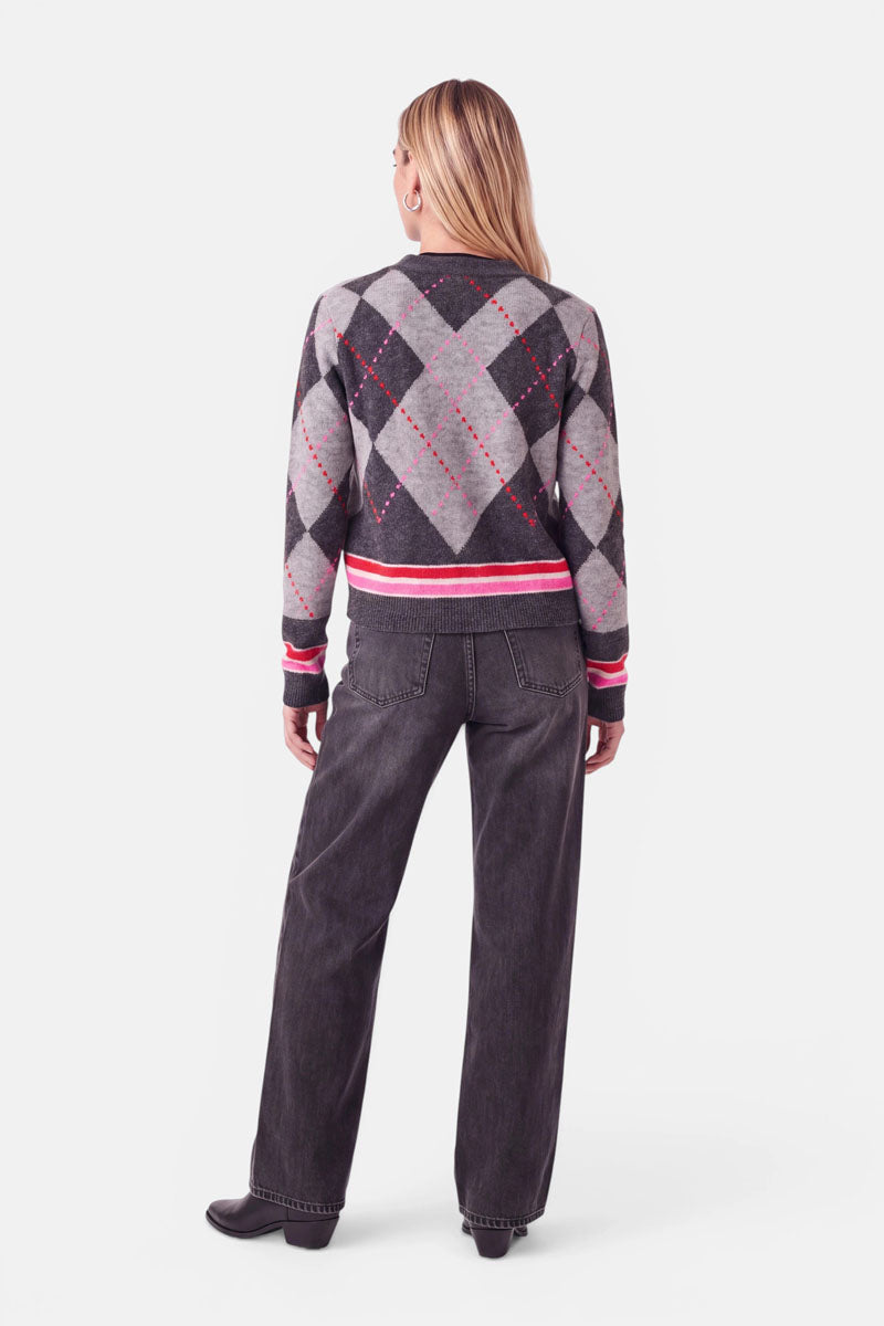 Eine Person mit glattem, blondem Haar trägt eine dunkelgraue Strickjacke mit einem Argyle-Muster aus roten und hellgrauen Rauten, dazu eine lockere dunkle Jeans und schwarze Schuhe; sie steht mit dem Rücken zur Kamera vor einem schlichten weißen Hintergrund.