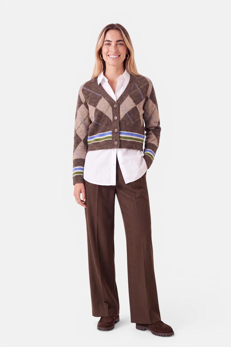 Eine Person steht lächelnd da und trägt eine braune Strickjacke mit Argyle-Muster und blauen und grünen Streifen, darunter ein weißes Hemd mit Knöpfen, eine braune Hose mit weitem Bein und braune Schuhe. Sie hat langes Haar und steht vor einem schlichten weißen Hintergrund.