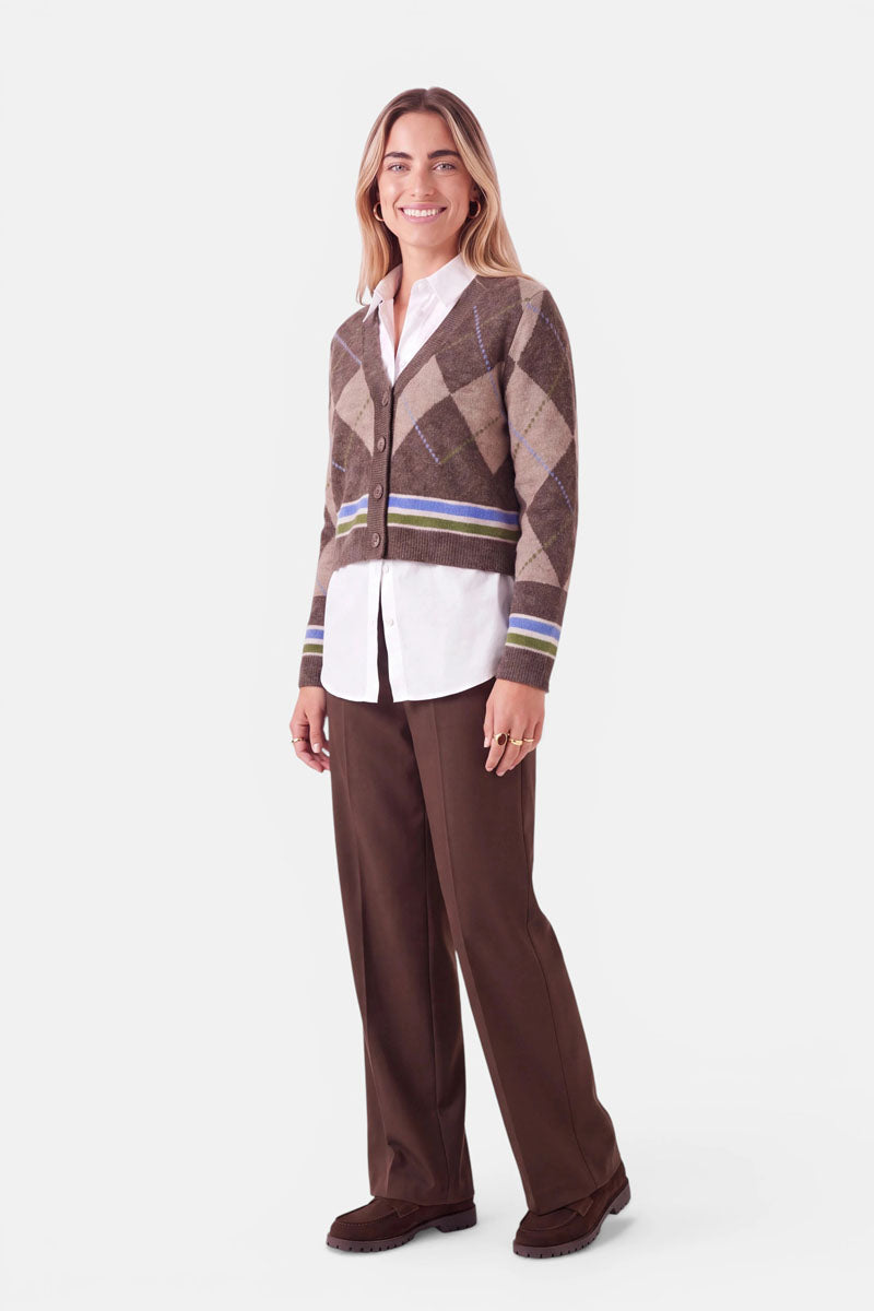 Eine Person mit langen Haaren lächelt vor einem schlichten weißen Hintergrund. Sie trägt eine braune Strickjacke mit Merinogewebe und einem Argyle-Muster aus grünen und blauen Streifen, darunter ein weißes Hemd mit Kragen, eine braune Hose mit weitem Bein und braune Schuhe. Die Hände liegen an den Seiten.