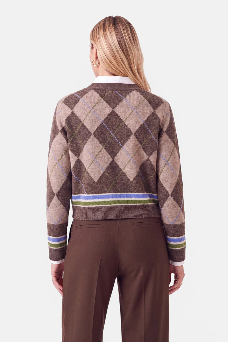 Eine Frau mit langen, glatten blonden Haaren trägt eine braun-beige Strickjacke mit Argyle-Muster und grünen sowie blauen Streifen am Saum und an den Ärmelbündchen, dazu eine braune Hose. Sie steht mit dem Rücken zur Kamera vor einem schlichten, hellen Hintergrund.