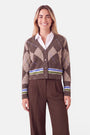 Eine Frau lächelt. Sie trägt ein weißes Hemd mit Kragen unter einer braunen Strickjacke, die am Saum und an den Ärmelbündchen blaue und grüne Streifen sowie ein Argyle-Muster aufweist. Sie hat langes Haar, goldene Creolen und eine braune Hose. Der Hintergrund ist schlicht weiß.