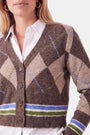 Eine Frau trägt eine braune Strickjacke mit Argyle-Muster in Beige, Blau und Grün über einem weißen Hemd mit Kragen. Die Strickjacke hat vier Knöpfe und horizontale blaue und grüne Streifen am Saum, was ihr einen klassischen Look verleiht, der durch einen Anteil an Merinowolle für zusätzliche Weichheit abgerundet wird. Sie hat helle Haut, blonde Haare und goldene Creolen.
