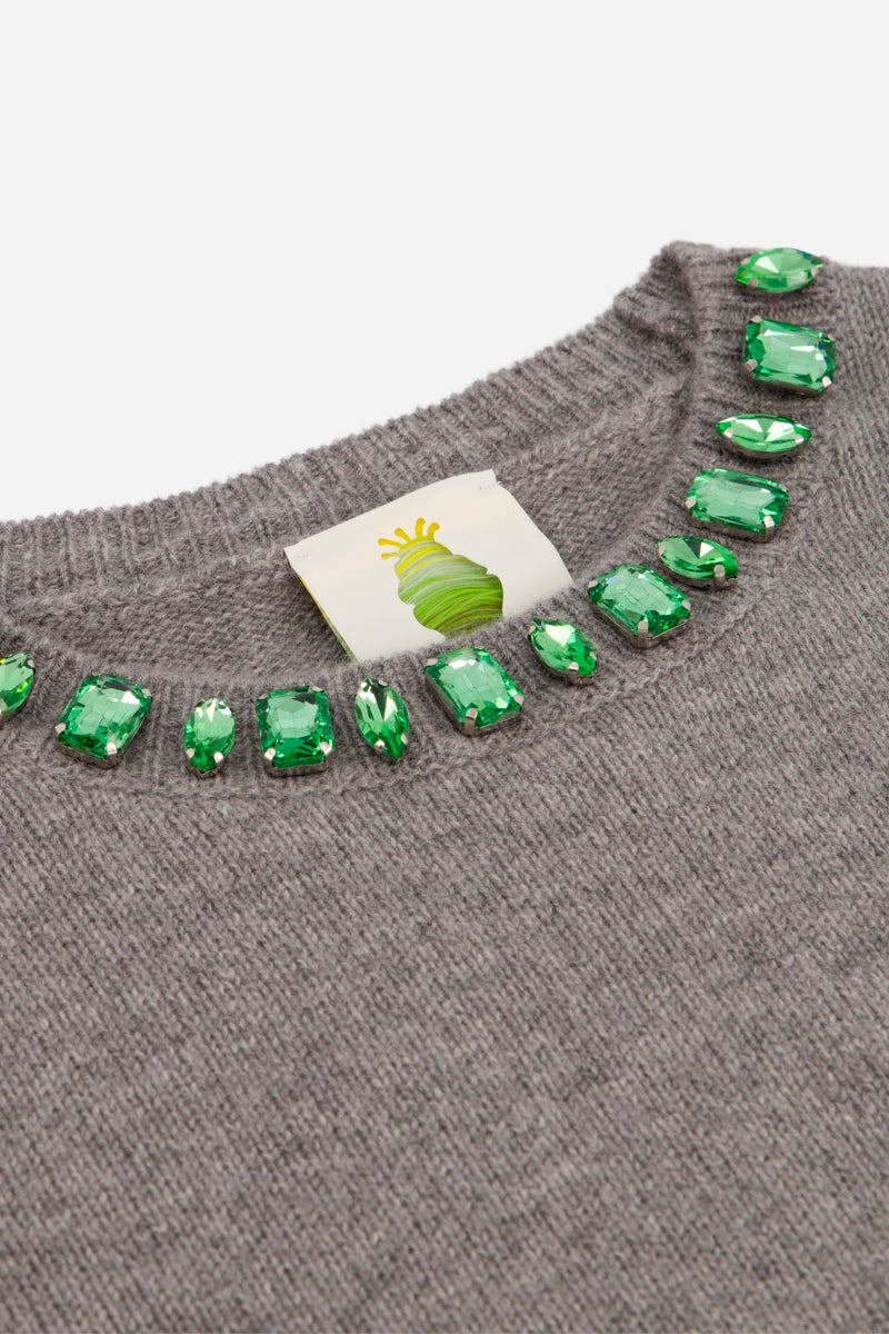 Ein grauer Strickpullover aus Wolle und Kaschmir mit einer Reihe smaragdgrüner, rechteckiger Kristalle, die den Rundhalsausschnitt säumen. Auf dem Innenetikett ist das Logo einer stilisierten grünen Krone abgebildet.