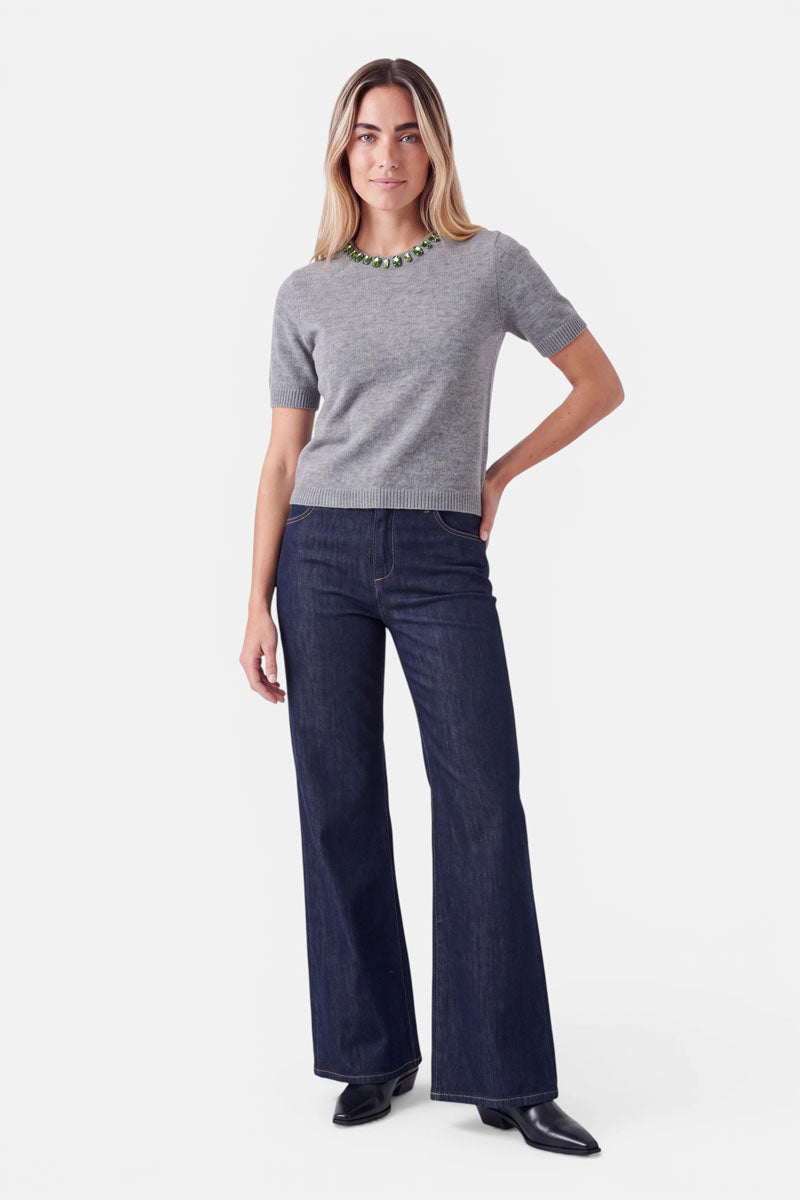 Eine Frau trägt einen grauen, kurzärmeligen Pullover aus einer Woll-Kaschmir-Mischung mit grünem, mit Perlen verziertem Rundhalsausschnitt, dazu eine dunkelblaue, weit geschnittene Jeans und schwarze Stiefel.