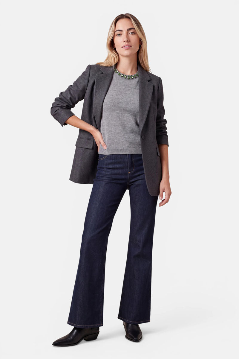 Eine Frau präsentiert einen grauen Blazer über einem passenden Pullover aus einer Woll-Kaschmir-Mischung und dunkelblauen Jeans. Sie trägt eine auffällige grüne Halskette und schwarze Stiefel.