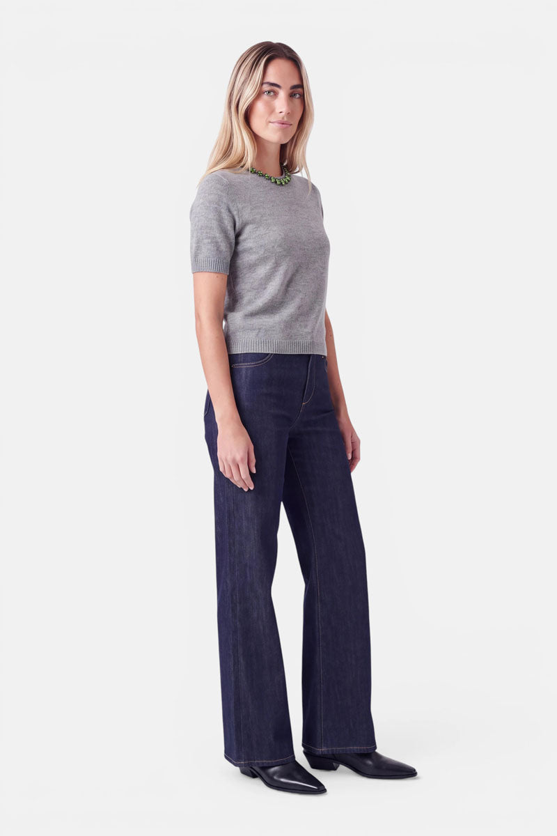 Eine Frau steht da und trägt einen grauen, kurzärmeligen Pullover aus einer Woll-Kaschmir-Mischung, dunkelblaue Jeans mit weitem Bein und schwarze Stiefel; das Outfit wird durch eine auffällige grüne Perlenkette mit Ziersteinen abgerundet.