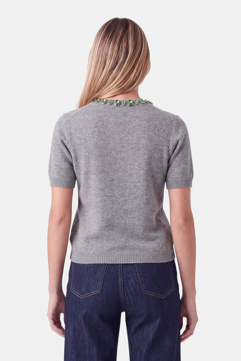Eine Frau, von hinten gesehen, die einen grauen Pullover mit kurzen Ärmeln und Rundhalsausschnitt sowie einem grünen, mit Edelsteinen besetzten Kragen trägt, dazu eine dunkle Jeans.