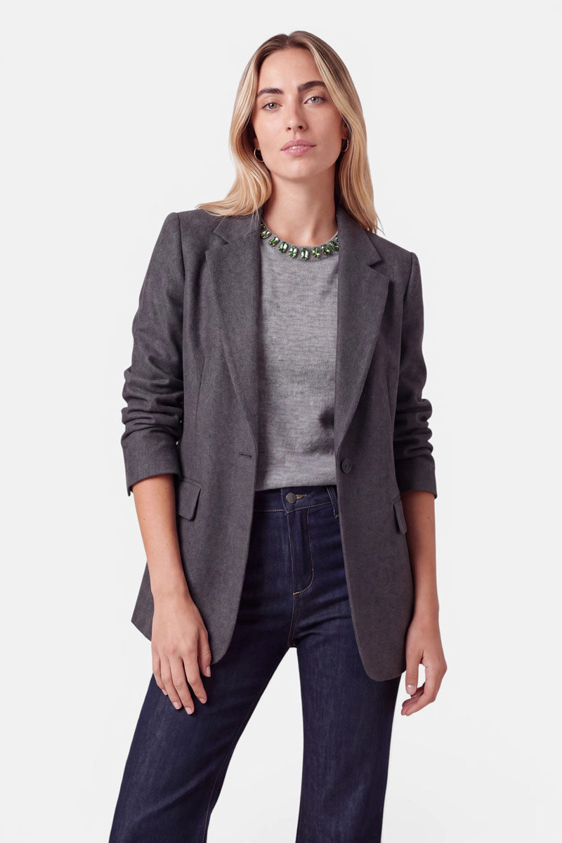 Eine Frau trägt einen grauen Blazer über einem grauen Pullover aus einer Woll-Kaschmir-Mischung und dunkelblauen Jeans. Als Accessoire trägt sie eine auffällige Halskette mit großen, grünen, facettierten Steinen.