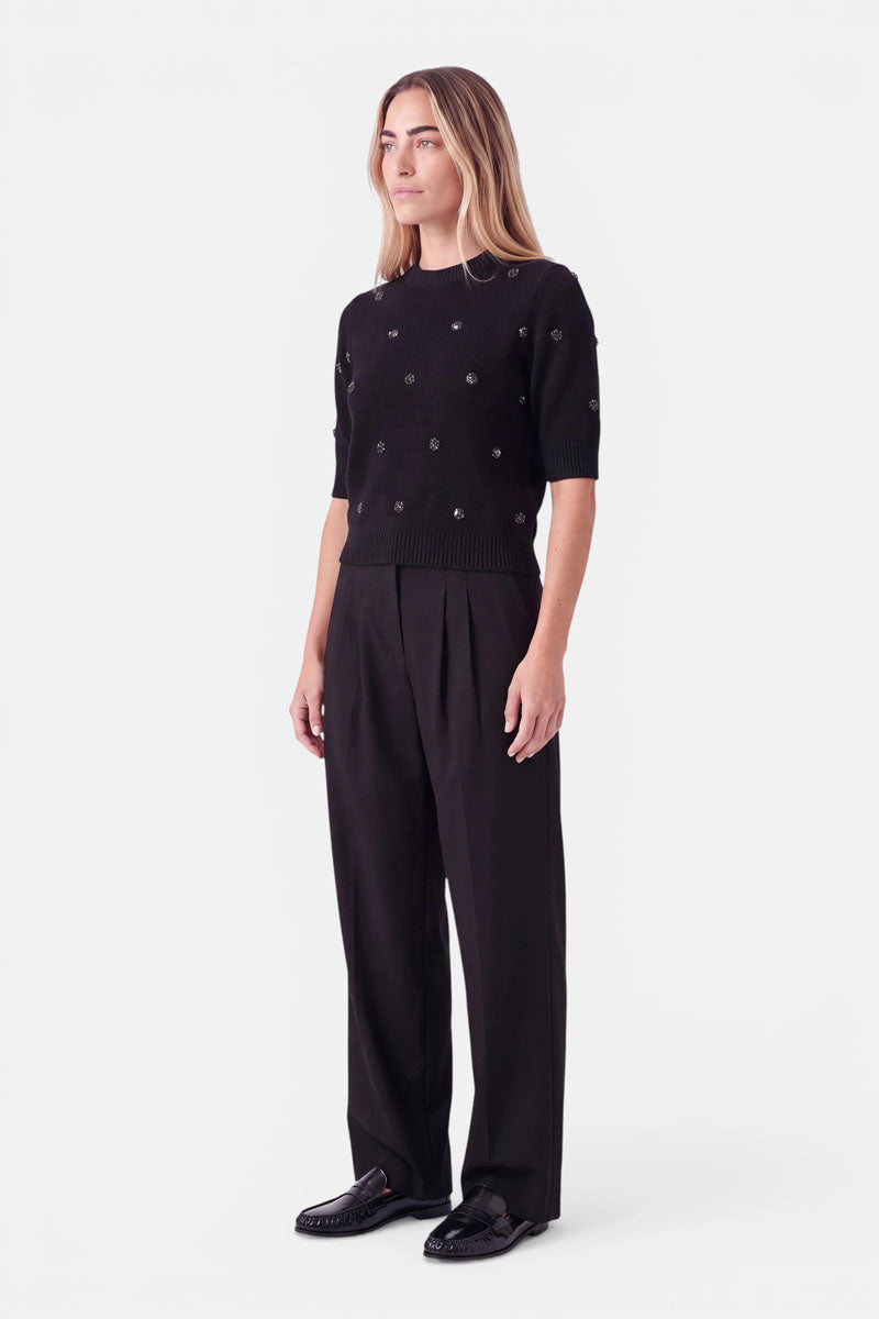 Eine Frau, die einen schwarzen, kurzärmeligen Pullover aus einer Woll-Alpaka-Mischung trägt, der mit kleinen runden Nieten verziert ist, sowie eine locker sitzende schwarze Hose. Dazu trägt sie schwarze Loafer und steht vor einem weißen Hintergrund.