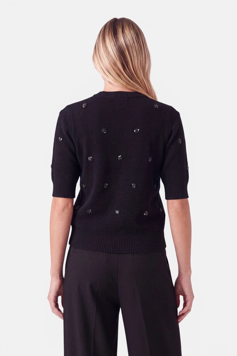 Rückansicht einer Person, die einen schwarzen, kurzärmeligen Pullover aus einer Woll-Alpaka-Mischung trägt, der mit vereinzelten, dunklen, runden Perlen verziert ist. Der Pullover hat einen gerippten Halsausschnitt, gerippte Ärmelbündchen und einen gerippten Saum.
