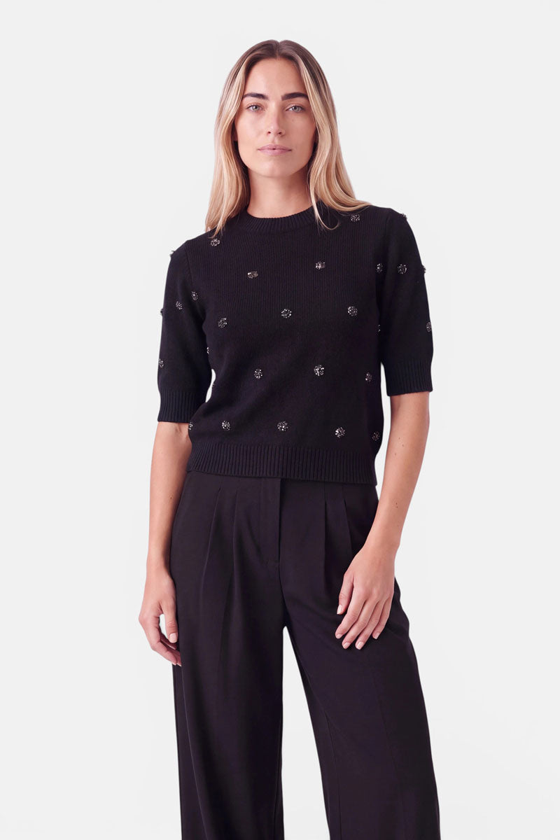 Eine Frau trägt einen schwarzen, kurzärmeligen Strickpullover aus einer Woll-Alpaka-Mischung, der mit vereinzelten Blumenakzenten aus Kristall verziert ist, dazu eine schwarze Hose.
