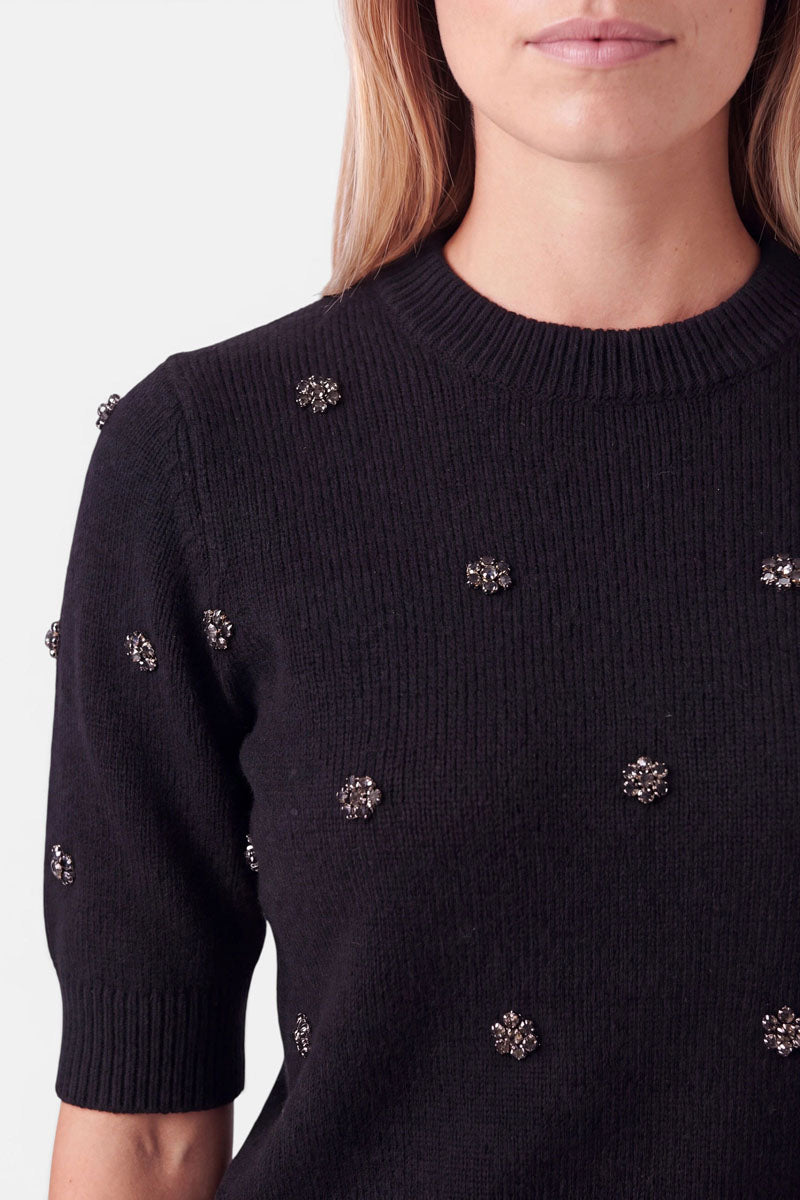 Eine Frau, die einen Strickpullover aus einer Woll-Alpaka-Mischung trägt, der mit kleinen, dekorativen, kristallartigen Blumenverzierungen versehen ist, die über Brust und Ärmel verteilt sind.