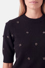 Eine Frau, die einen Strickpullover aus einer Woll-Alpaka-Mischung trägt, der mit kleinen, dekorativen, kristallartigen Blumenverzierungen versehen ist, die über Brust und Ärmel verteilt sind.