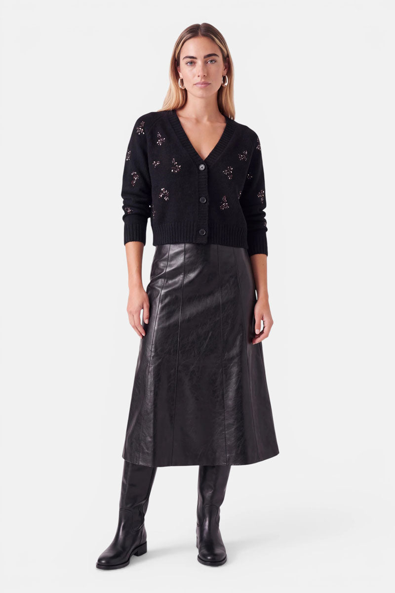 Eine Frau präsentiert eine schwarze Strickjacke mit V-Ausschnitt und Knopfleiste aus einer Woll-Alpaka-Mischung, die mit kleinen Blumenverzierungen und Strasssteinen verziert ist, dazu trägt sie einen schwarzen Midirock und kniehohe Stiefel.