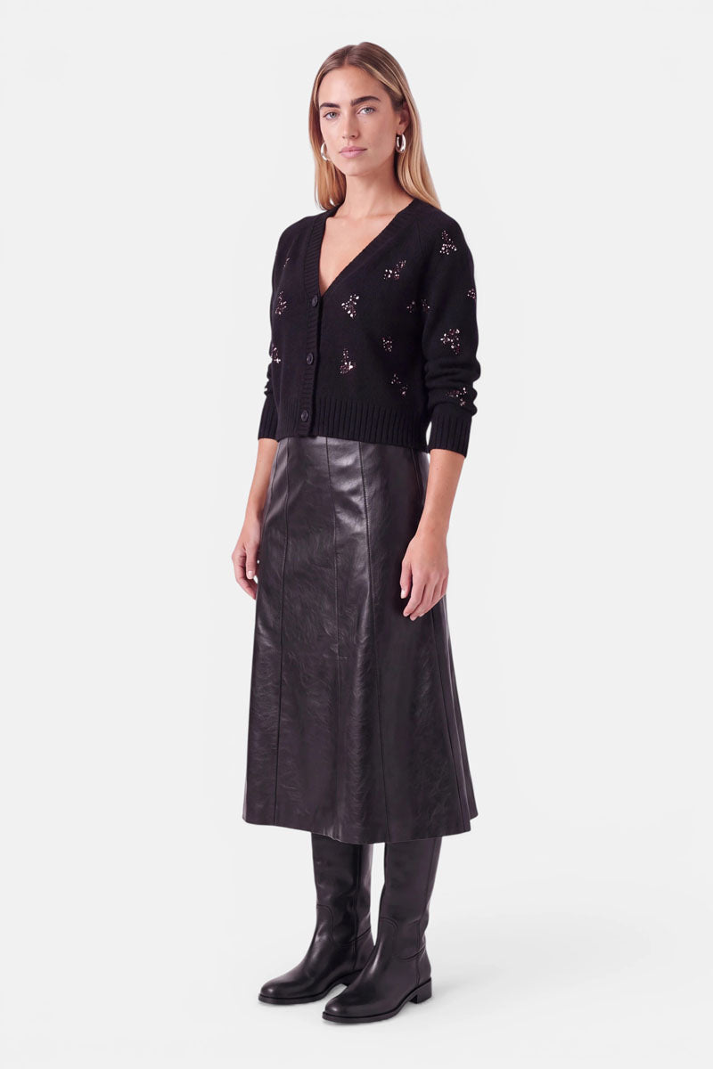 Eine Frau trägt eine schwarze Strickjacke mit V-Ausschnitt aus einer Woll-Alpaka-Mischung und kleinen, ausgeschnittenen Blumendetails, einen langen schwarzen A-Linien-Rock und schwarze kniehohe Stiefel.