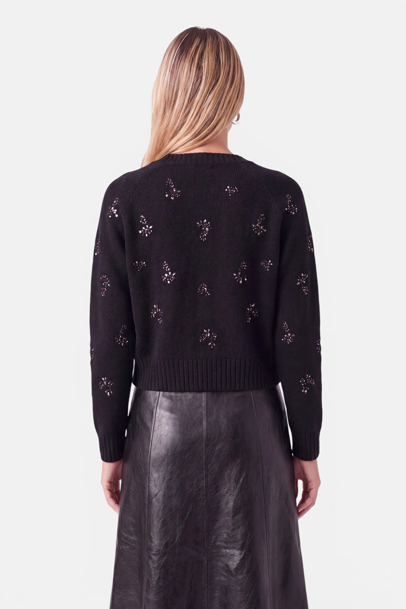 Eine Person, die einen schwarzen, langärmeligen Pullover aus einer Woll-Alpaka-Mischung trägt, der mit kleinen, funkelnden Strassblumen verziert ist, dazu einen schwarzen Rock. Der Pullover hat einen gerippten Saum und gerippte Bündchen.