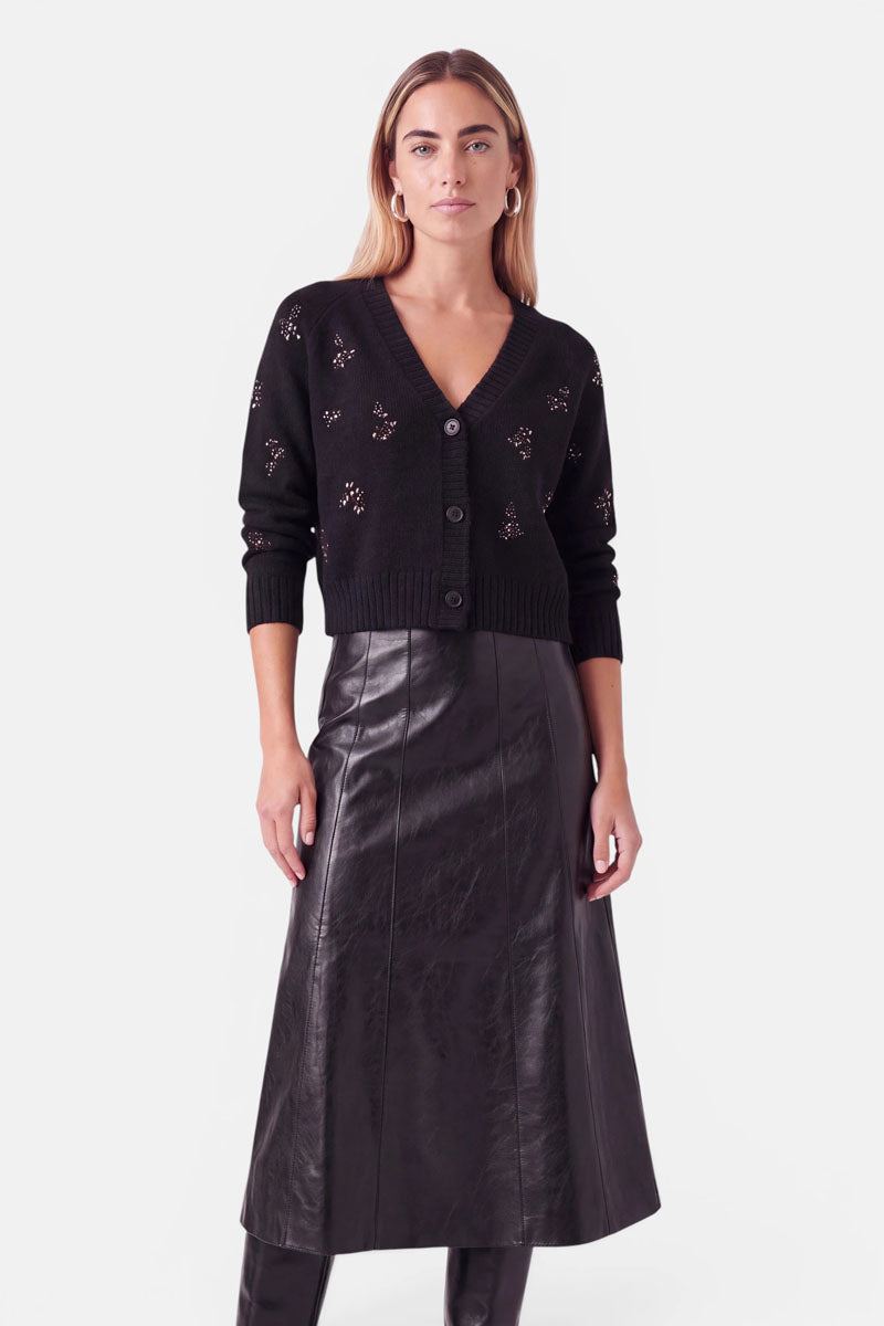 Eine Frau steht mit dem Blick nach vorne und trägt eine schwarze Strickjacke mit V-Ausschnitt und Knöpfen sowie kleinen Verzierungen, einen schwarzen Midirock aus veganem Leder mit einem dezenten Reißverschlussdetail und schwarze kniehohe Stiefel. Sie hat hellbraunes Haar, trägt Creolen und posiert vor einem schlichten weißen Hintergrund.