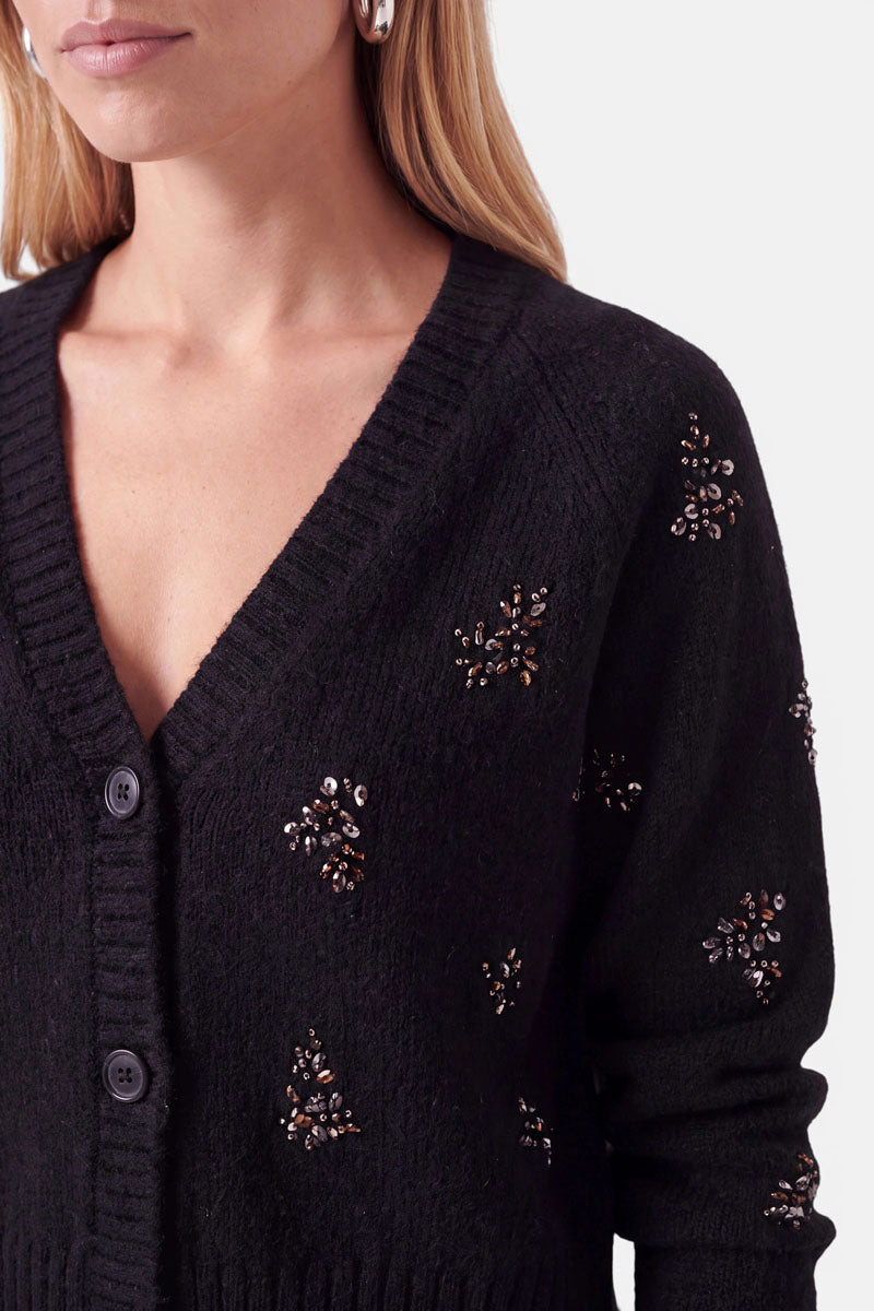 Eine Person trägt eine schwarze Strickjacke aus einer Woll-Alpaka-Mischung mit Knöpfen und V-Ausschnitt, die mit vereinzelten Gruppen dunkler, funkelnder Strassperlen verziert ist.