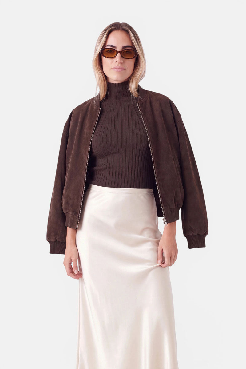 Eine Frau, die eine braune, ovale Sonnenbrille, einen braunen, gerippten Rollkragenpullover und eine braune Bomberjacke über einem cremefarbenen Midirock trägt. Sie blickt nach vorne und hat die Hände nahe am Saum des Rocks aufgelegt.
