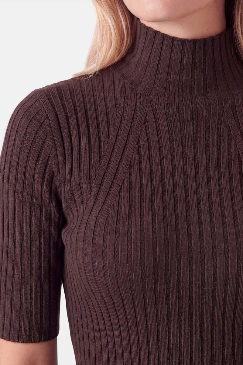 Eine Person trägt einen dunkelbraunen, gerippten Pullover mit Stehkragen und kurzen Ärmeln. Der Rippstrick-Schnitt weist dekorative vertikale Nähte entlang der Brust auf.