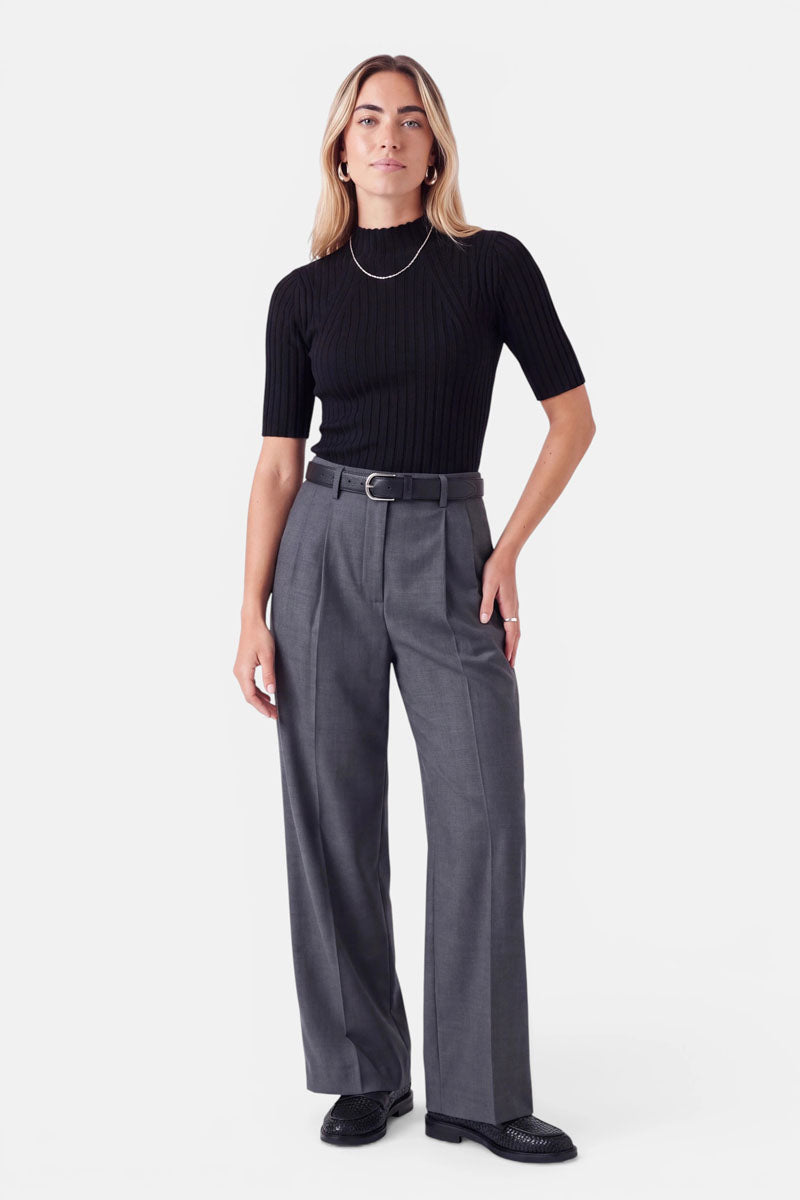 Eine Frau, die ein schwarzes, geripptes Kurzarm-Oberteil mit Stehkragen trägt, das in eine graue Hose mit weitem Bein und einem schwarzen Gürtel gesteckt ist. Sie trägt kleine Creolen, eine dünne Kette und schwarze Loafer.
