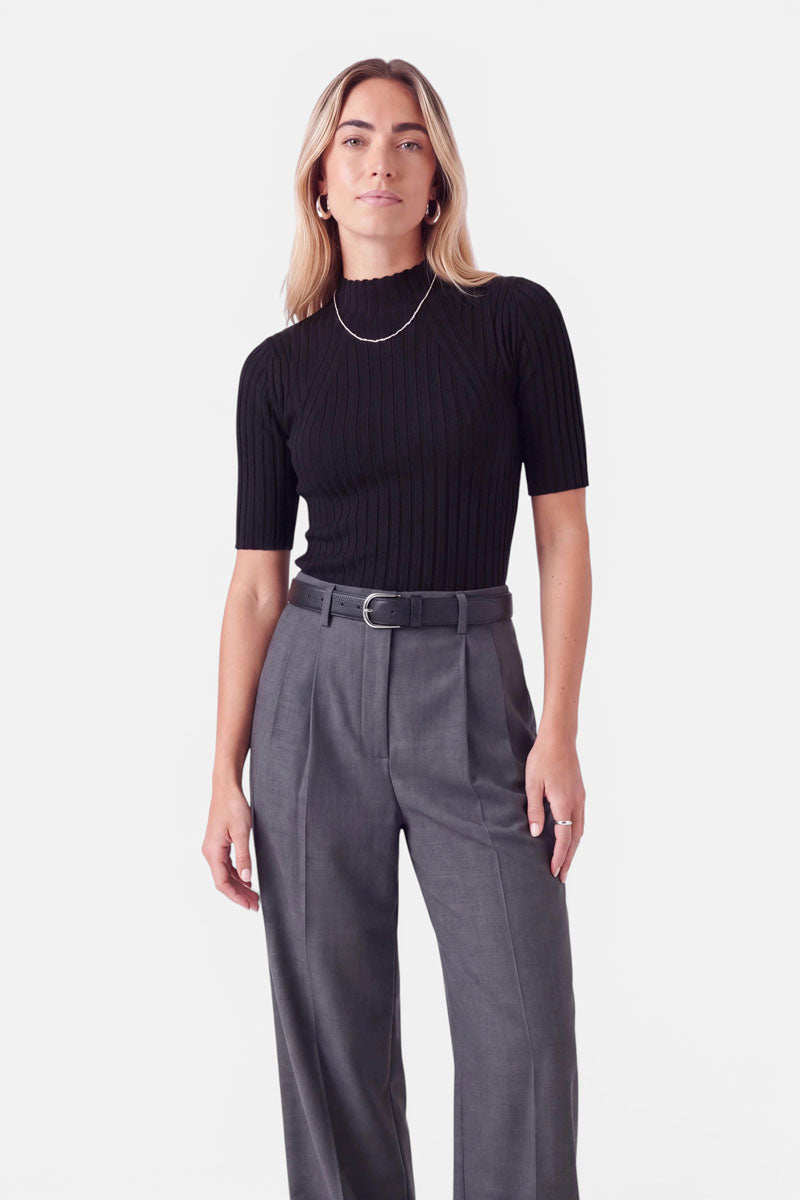 Eine Frau trägt ein schwarzes, kurzärmeliges, geripptes Oberteil mit Stehkragen, das sie in eine graue Plisseehose gesteckt hat. Als Accessoires trägt sie einen schmalen schwarzen Gürtel und eine silberne Kettenhalskette.