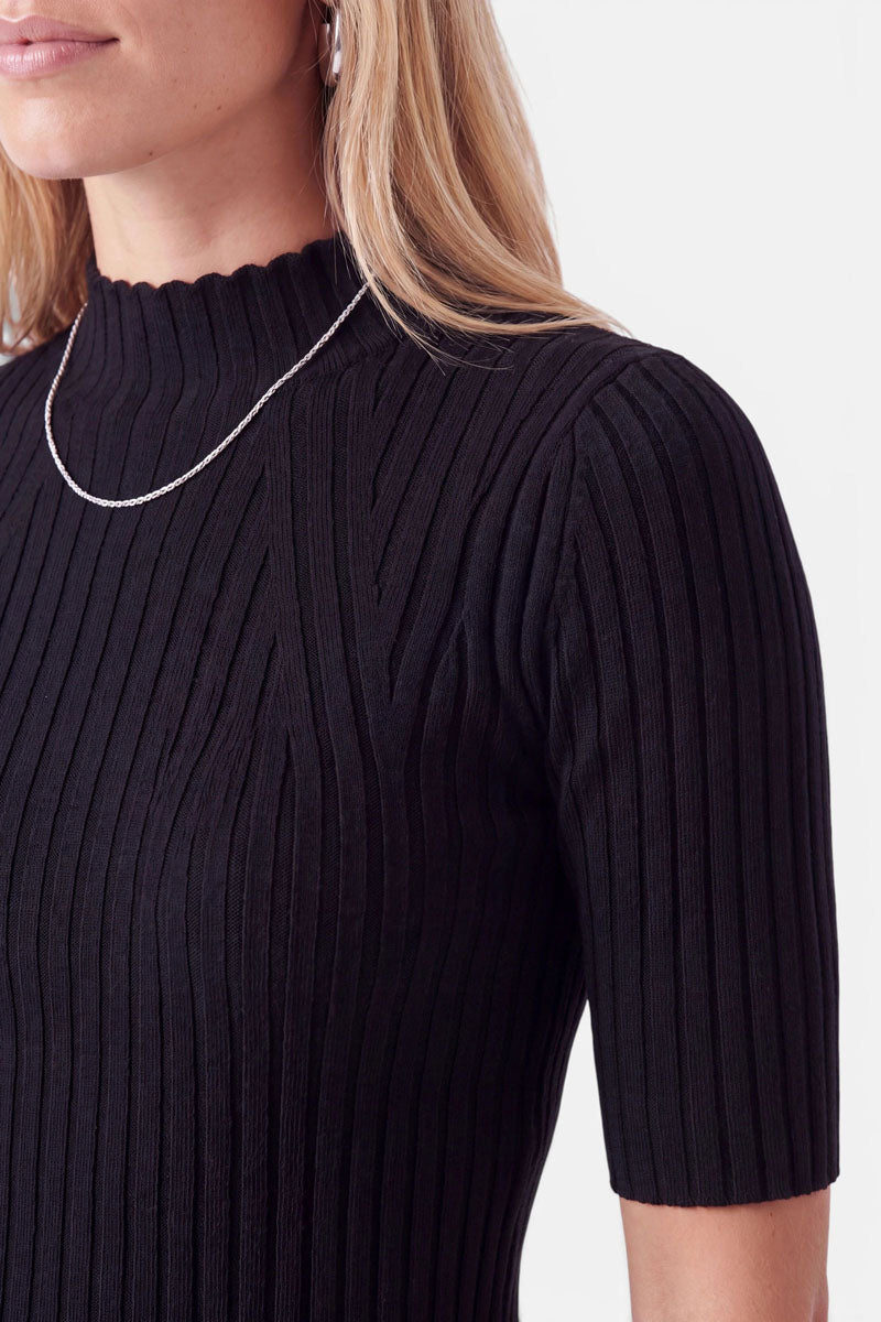 Eine Frau, die einen schwarzen, gerippten Pullover mit Rollkragen und kurzen Ärmeln trägt. Sie trägt dazu eine dünne silberne Kette und kleine silberne Creolen.