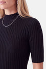 Eine Frau, die einen schwarzen, gerippten Pullover mit Rollkragen und kurzen Ärmeln trägt. Sie trägt dazu eine dünne silberne Kette und kleine silberne Creolen.