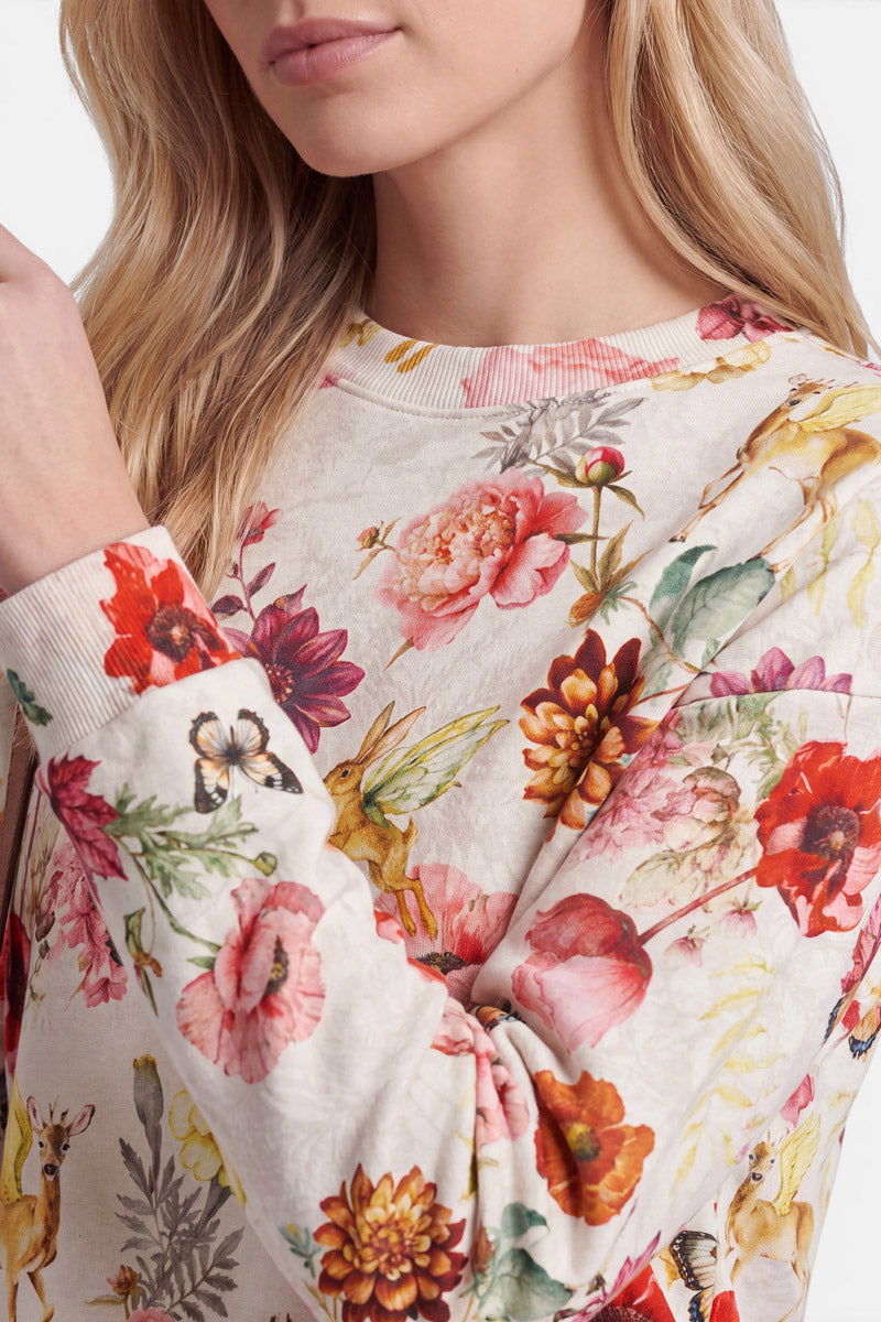 Eine Frau trägt einen cremefarbenen Sweater mit langen Ärmeln, der mit einem durchgehenden Print aus leuchtenden Blumenmotiven mit Rehen, Kaninchen und Schmetterlingen verziert ist. Ihr blondes Haar fällt ihr offen über die Schultern, und nur der untere Teil ihres Gesichts ist zu sehen. Sie hält ihren linken Arm hoch, wodurch ihr Mund teilweise verdeckt wird. Der weiche Baumwollstoff verleiht diesem verspielten Design zusätzlichen Tragekomfort.