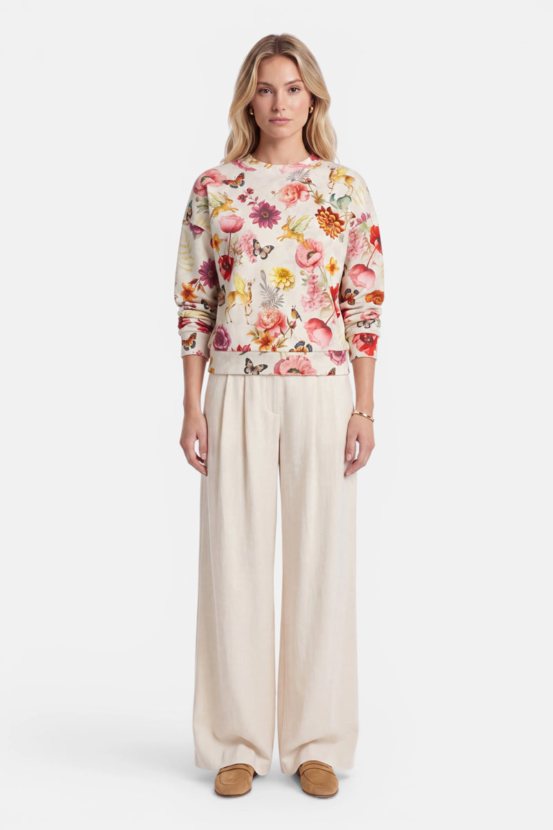 Eine Frau steht mit dem Blick nach vorne und trägt einen cremefarbenen Baumwoll-Sweater mit einem bunten Allover-Blumenprint, eine beige Hose mit weitem Bein und hellbraune Schuhe. Ihre Hände liegen an den Seiten, und sie hat leicht gewelltes Haar. Der Hintergrund ist schlicht weiß.