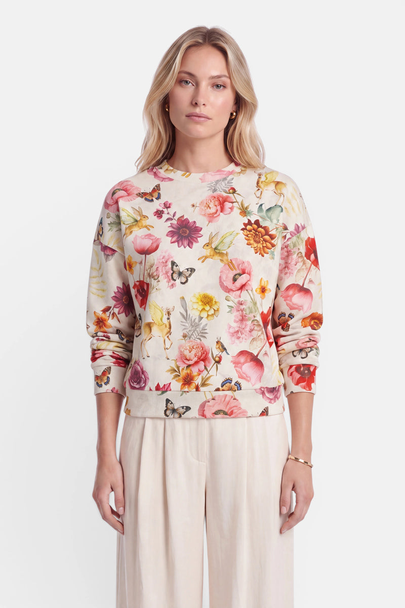 Eine Frau mit blonden Haaren trägt einen cremefarbenen Sweater mit einem durchgehenden Print aus bunten Blumen und Schmetterlingen. Sie steht mit neutralem Gesichtsausdruck nach vorne gerichtet da. Außerdem trägt sie eine hellbeige Hose, ein goldenes Armband und schlichte Ohrringe. Der Hintergrund ist schlicht weiß.